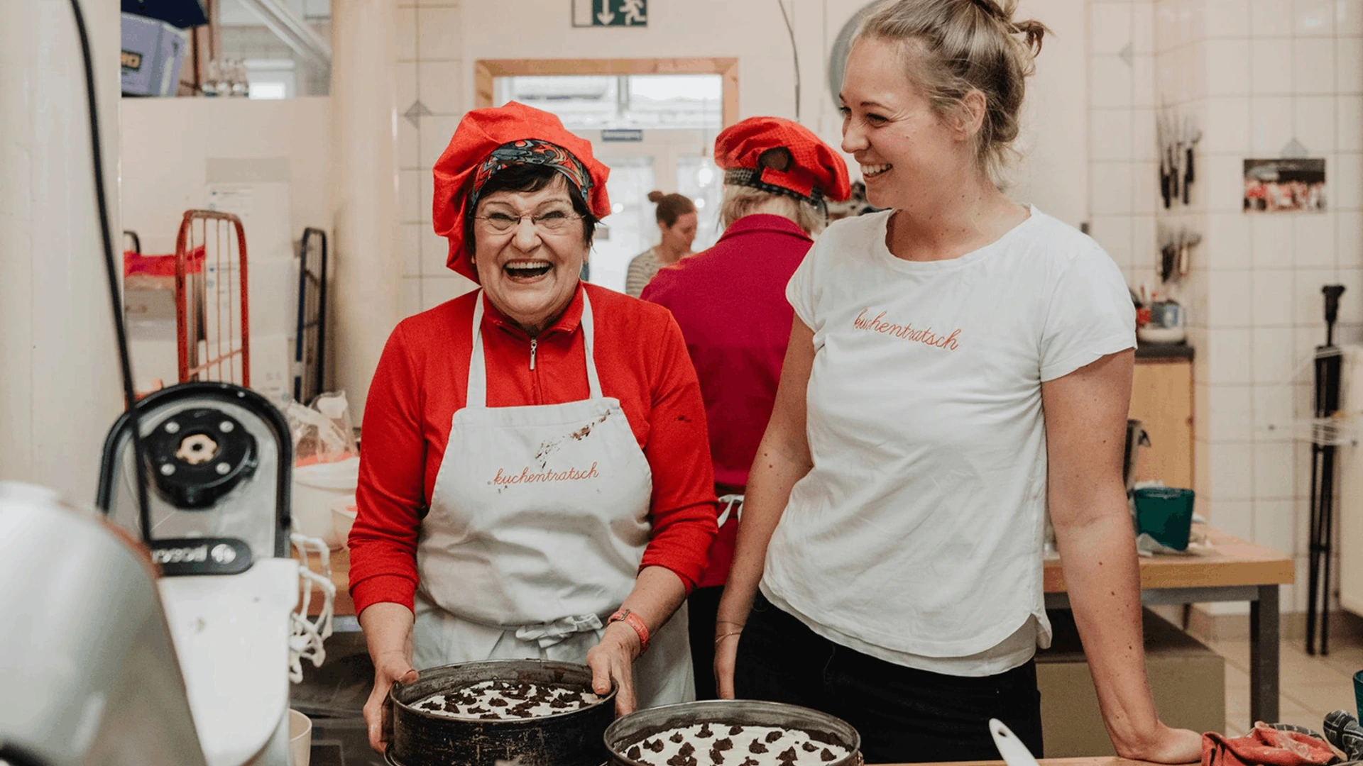 Zwei lächelnde Frauen in Küche mit kuchentratsch-Schürzen halten Kuchen, PALMBERG