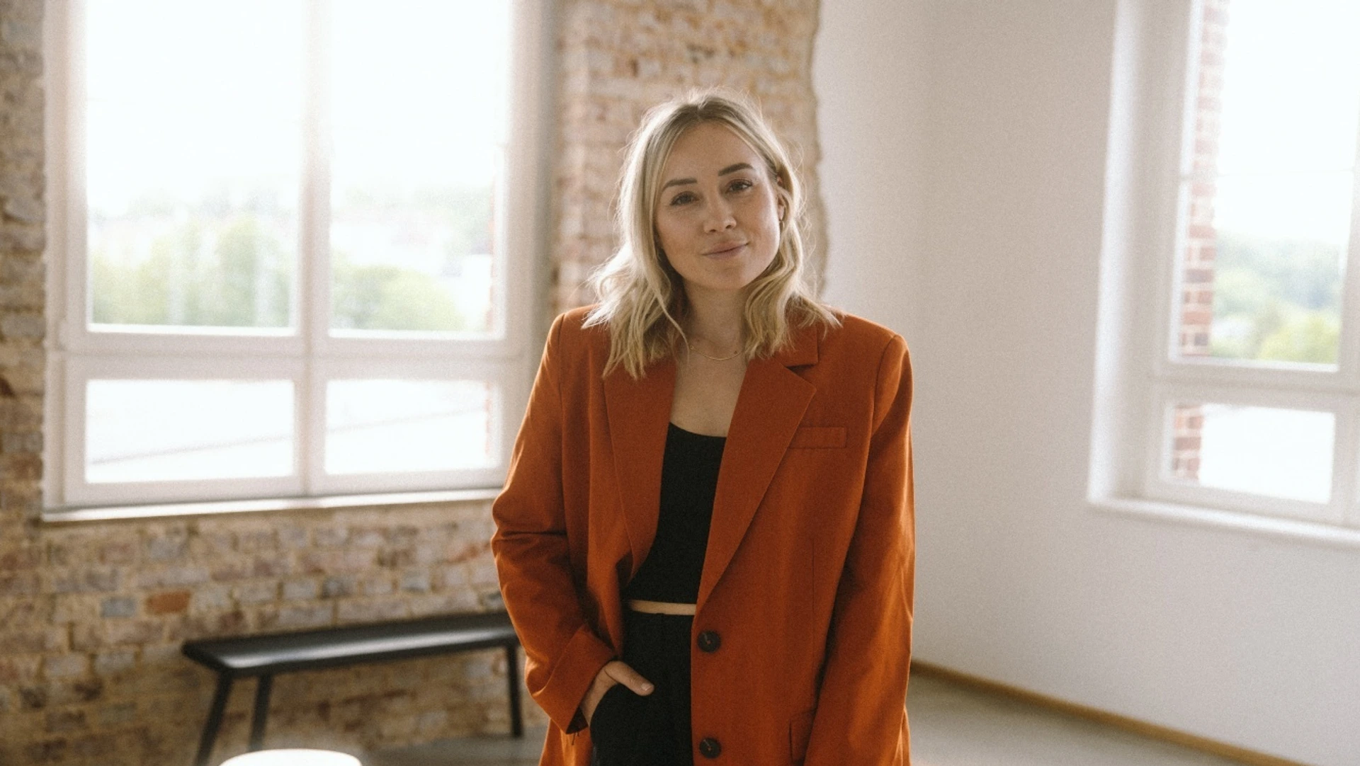 Frau in orangefarbener Jacke steht lächelnd im hellen Loft-Büro, im Hintergrund eine PALMBERG-Sitzbank.