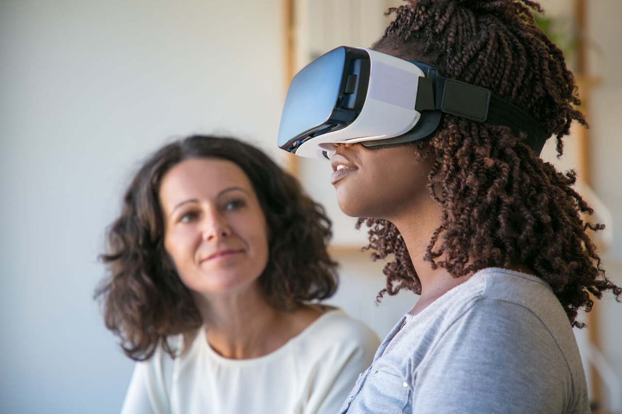 Zwei Frauen, die eine trägt eine VR-Brille und hat den Kopf leicht nach oben gerichtet, die andere Frau beobachtet sie.