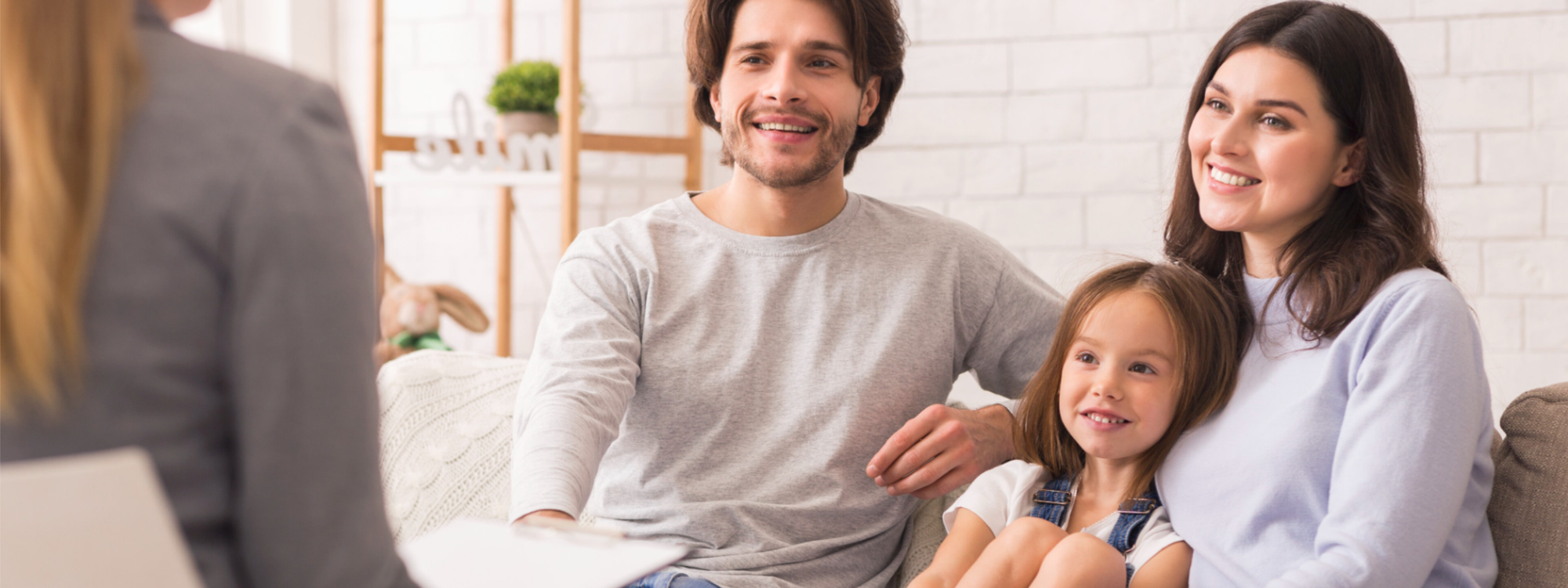 Eltern mit Kind sitzen lächelnd auf einem Sofa und sprechen mit einer Fachkraft im Beratungsgespräch