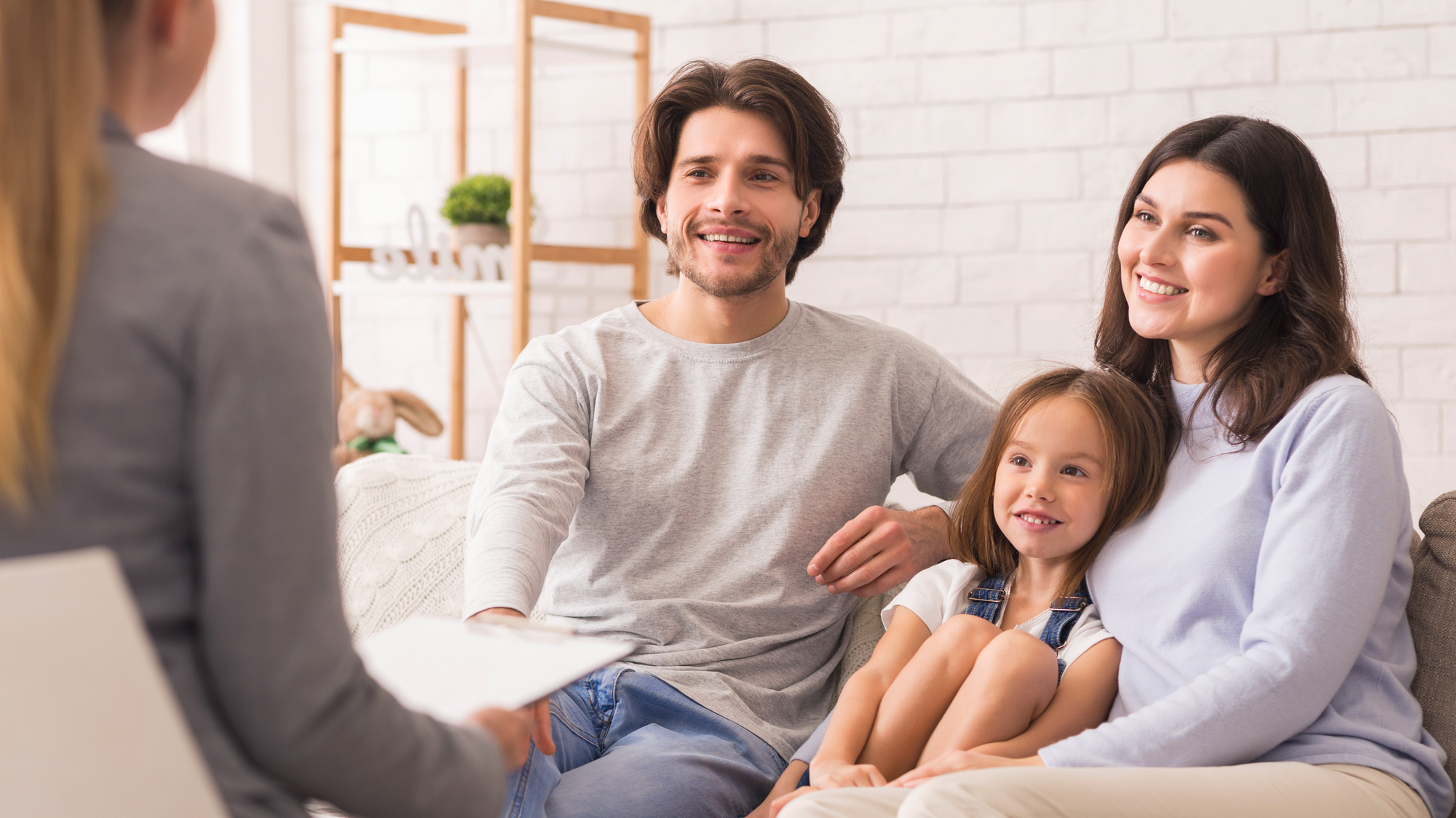 Eine Familie mit Vater, Mutter und Tochter sitzt lächelnd auf einem Sofa und spricht mit einer Beraterin oder Therapeutin.