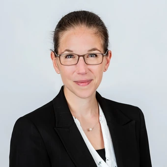 Foto zeigt eine junge, freundlich lächelnde Frau mit Brille, weißer Bluse und schwarzem Blaser.
