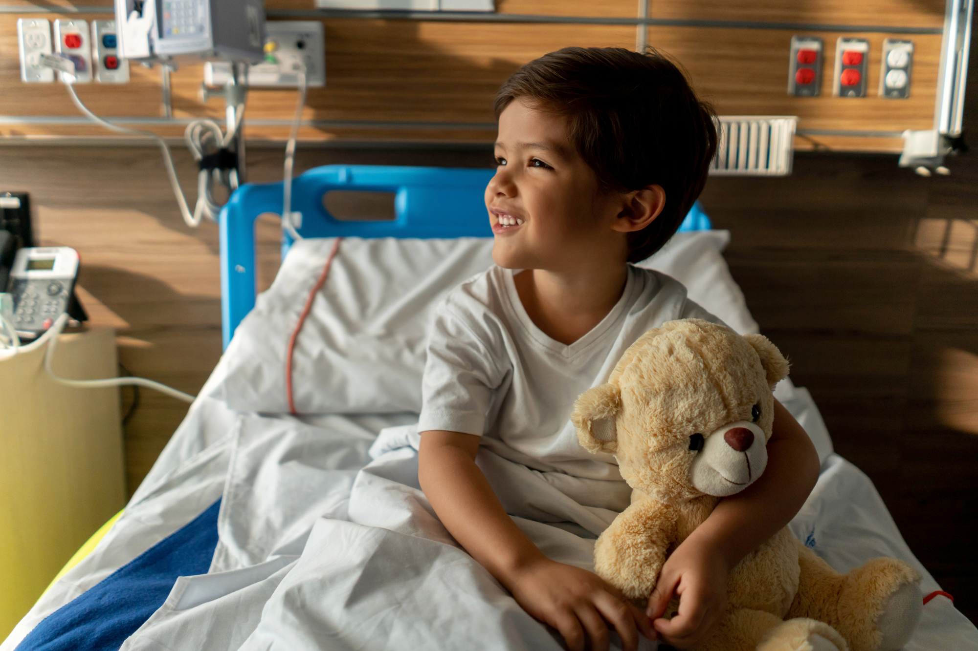 Cute boy sitting down on hospital bed hugging his teddy bear smiling