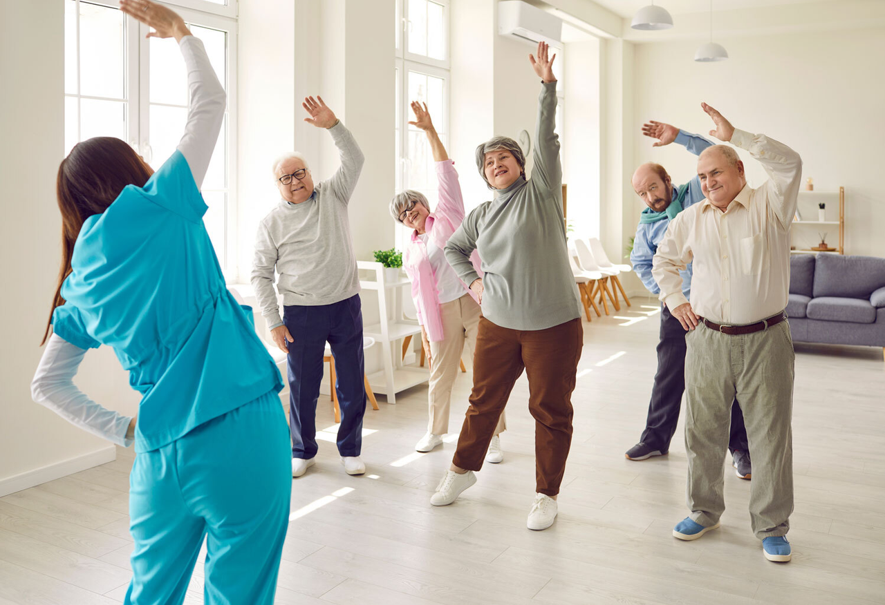 Eine Gruppe alter Menschen macht Gymnastik mit einer Gesundheitsfachkraft, diese ist nur von hinten zu sehen, die Senioren dehnen ihre Arme über den Kopf auf eine Seite