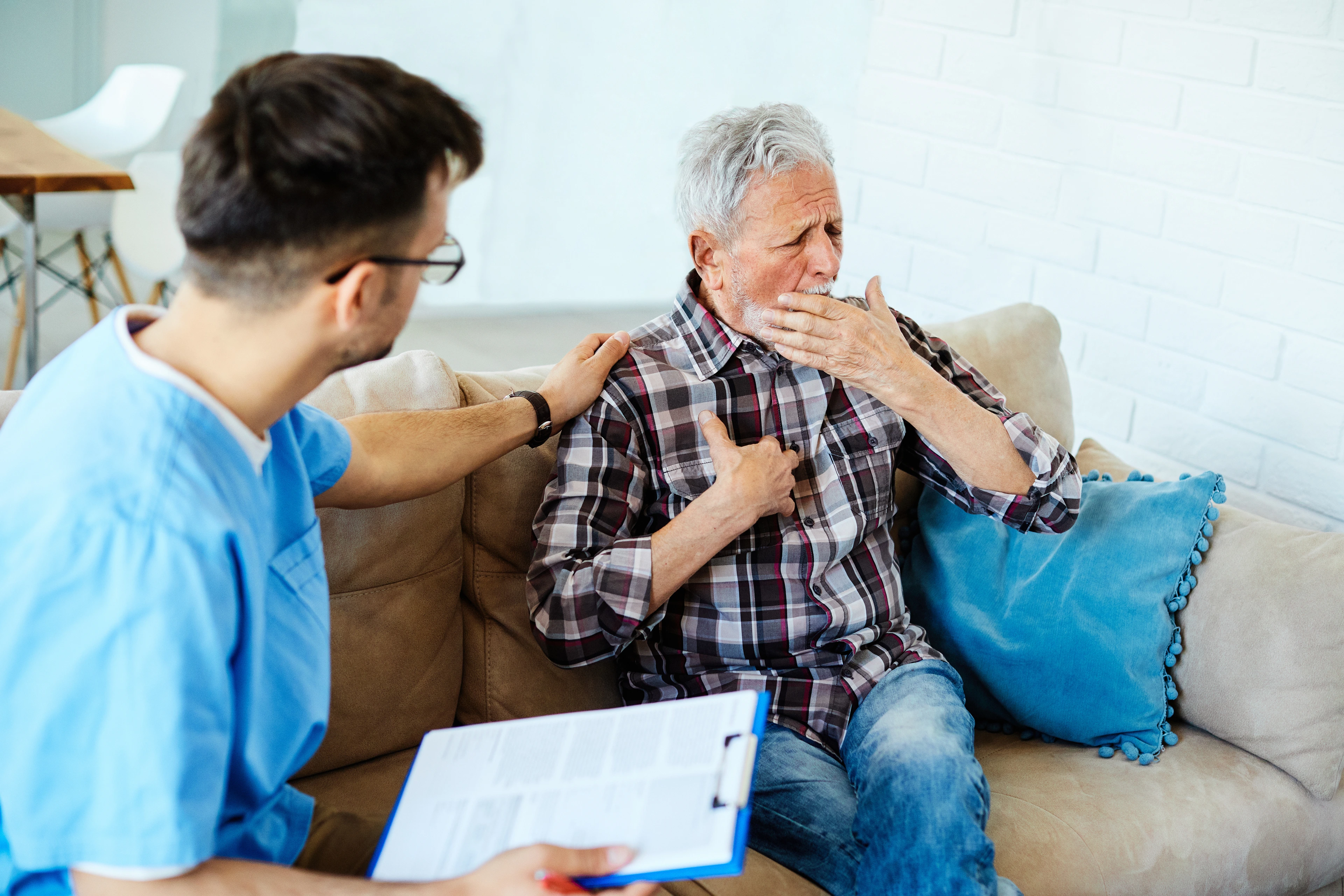 Doctor or nurse caregiver talking with senior man with chest pain and coughing problems on sofa at home or nursing home