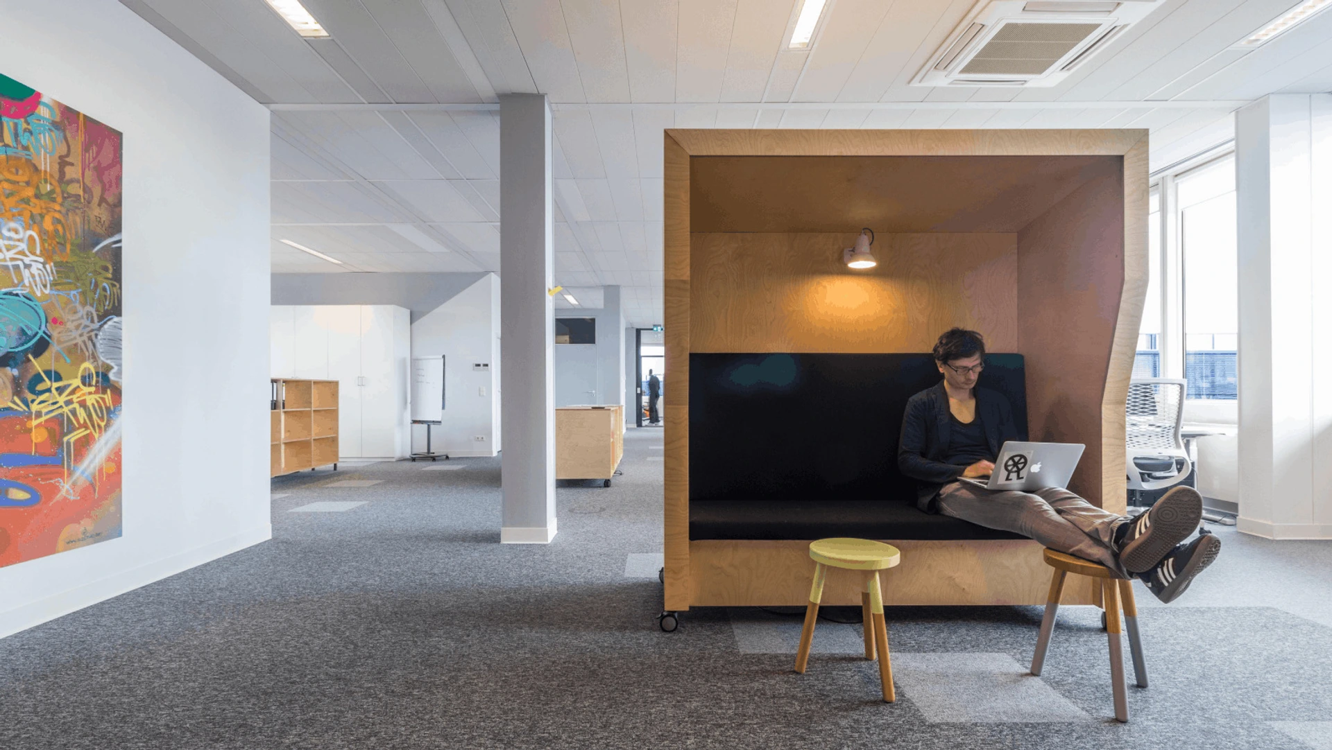 Person sitzt in einer hölzernen PALMBERG-Lounge, Beine auf Hocker, arbeitet am Laptop im offenen Büro