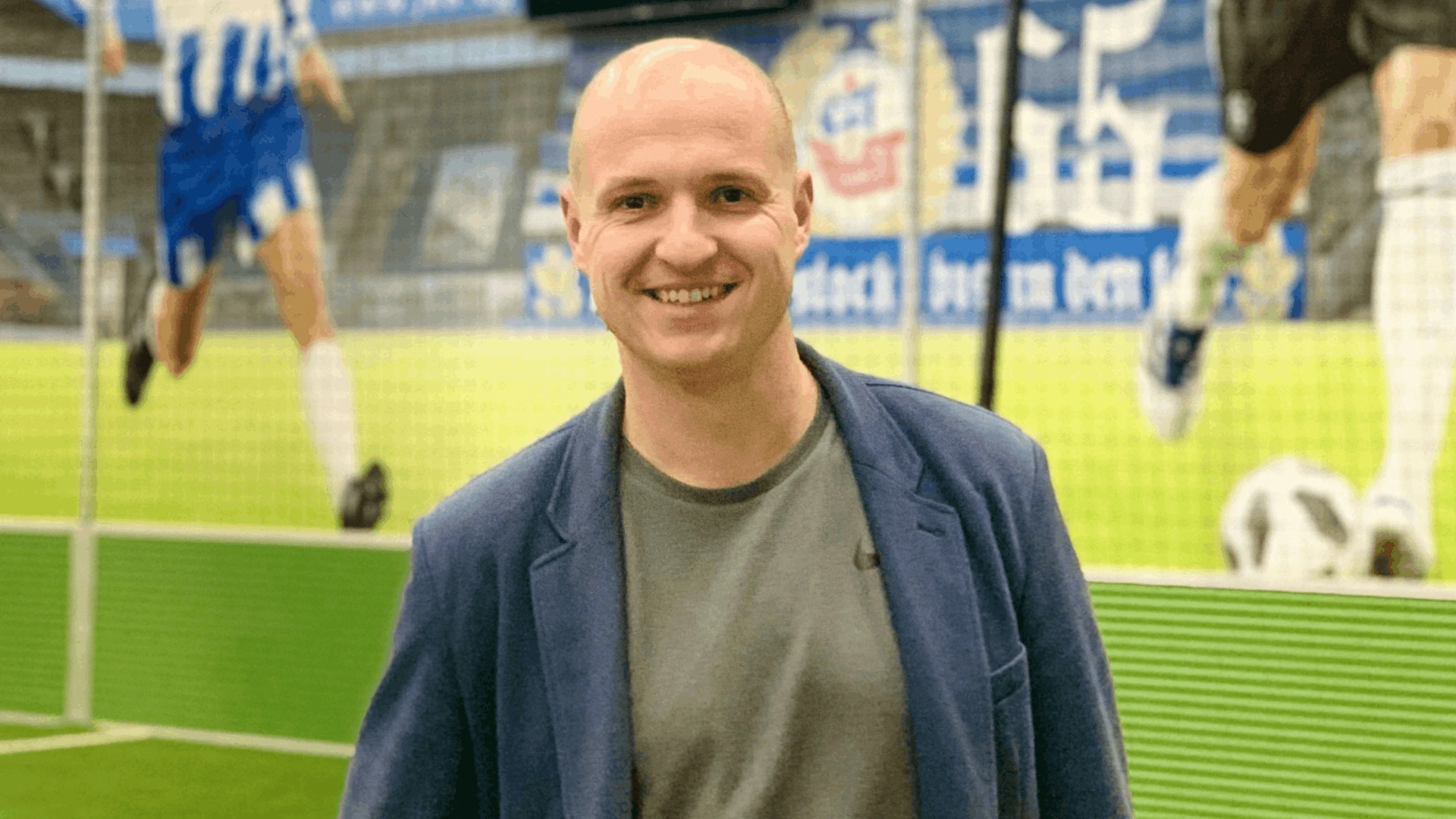 Lächelnder Mann steht vor Stadionwand mit Fußballspielern, PALMBERG.