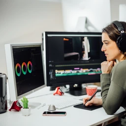 Profile view of Female designer sitting, editing video on a computer at her workplace. Modern business woman working concentrated in video design with a pen tablet in a studio. Freelance worker.