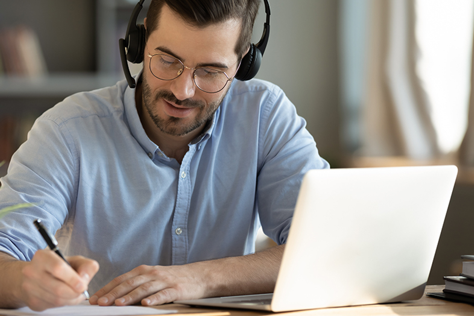 Man wearing a headset, working on a laptop and writing something down with pen and paper.