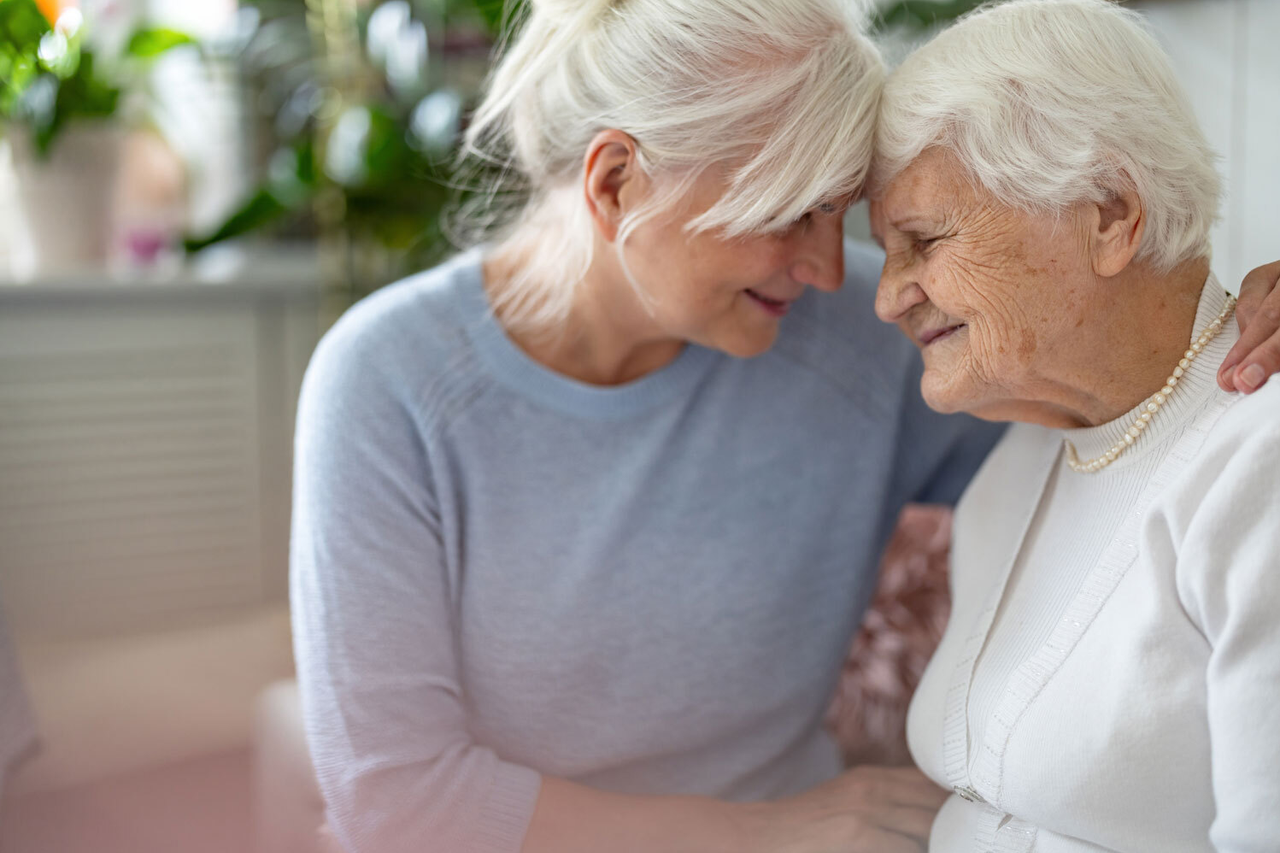 Eine Frau um die 60 drückt ihre Stirn an die Stirn einer Frau um die 80, sie umarmen sich