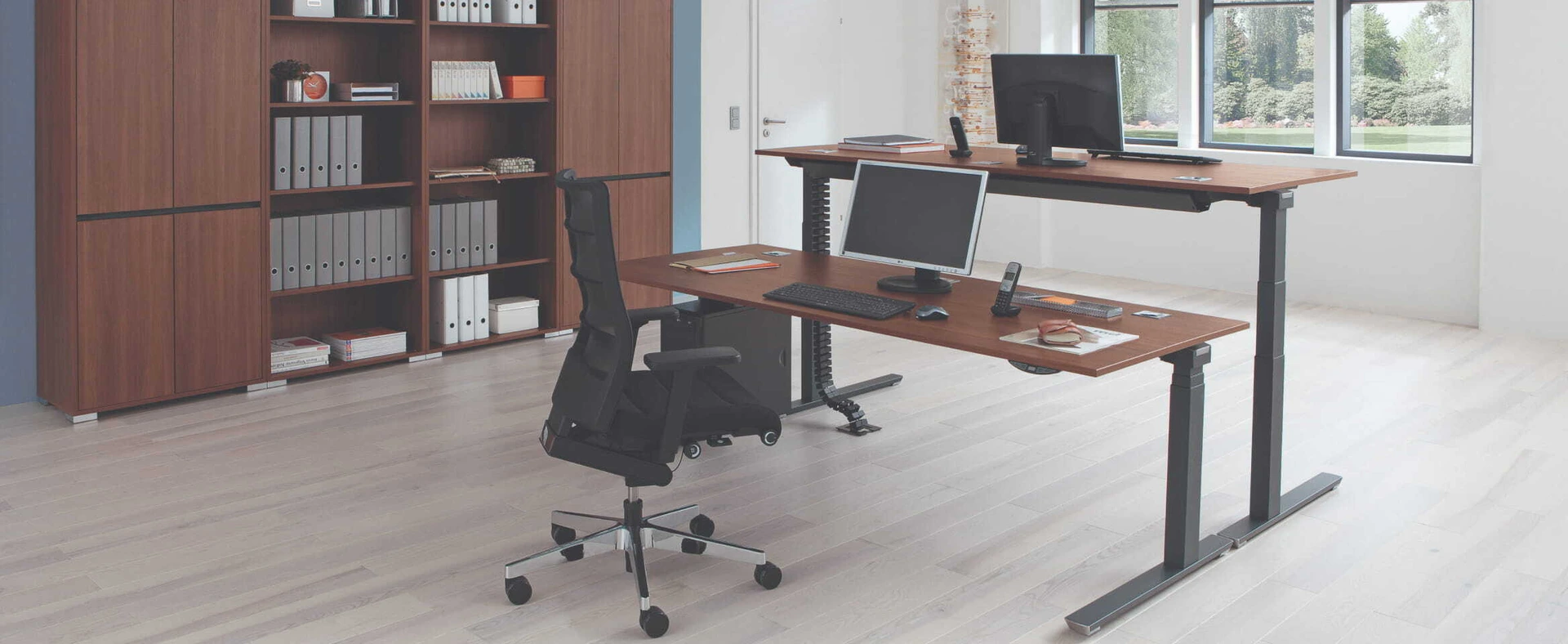 Zwei Tische in einem hellen Büro mit dunkler Möblierung. Einer der Tische ist für sitzendes Arbeiten eingestellt, der andere für Steh-Arbeit.